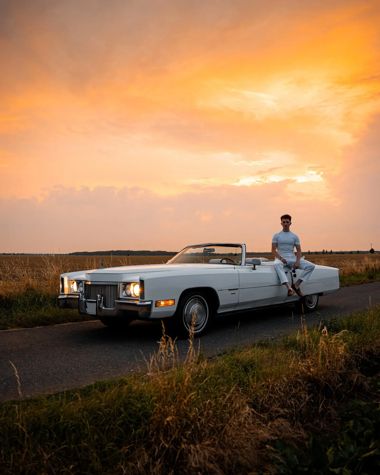 Muž sedí na zádi bílého Cadillacu Eldorado na silnici pod oranžovou večerní oblohou